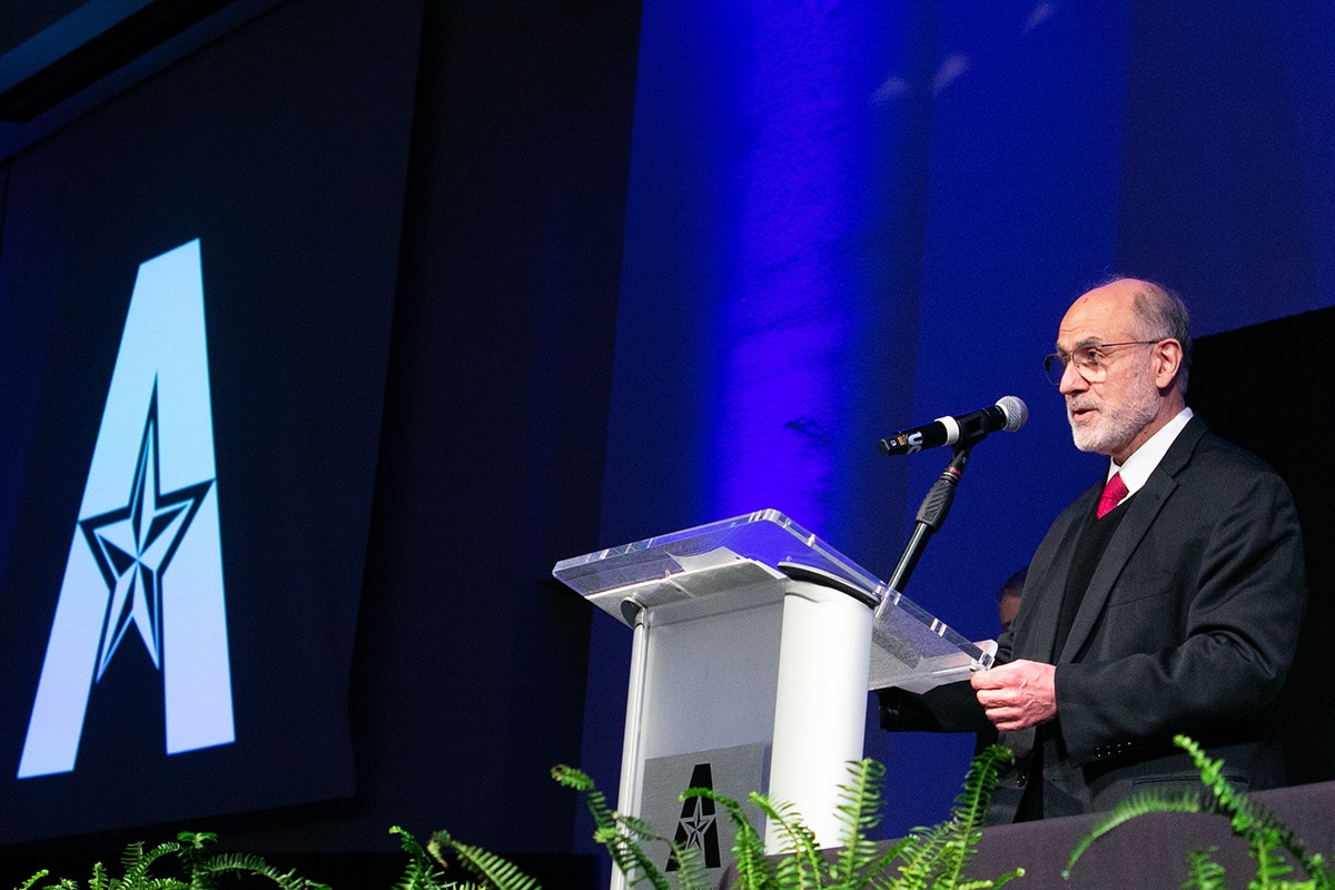 Man standing at a podium on a stage. Behind him on the wall is a large UTA logo - opens in a new tab