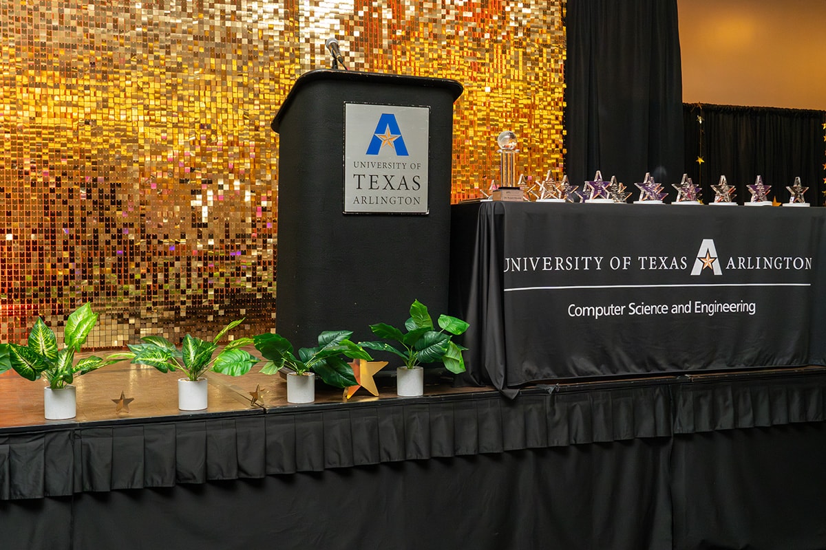 A stage at the University of Texas at Arlington with a podium, a table with star-shaped trophies, and a gold sequin backdrop - opens in a new tab